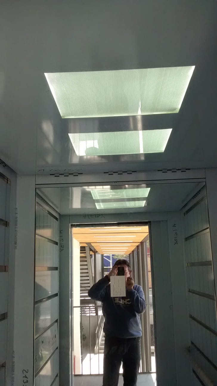 Cabina pequeña tipo montacargas con paredes metálicas grises, pasamanos de acero y piso antideslizante.