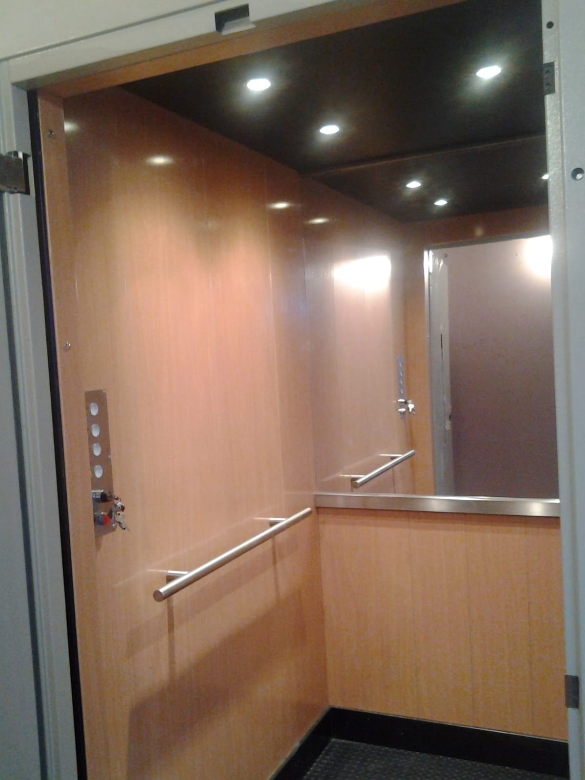 Cabina con acabado en madera clara, espejo al fondo, pasamanos metálico y panel de control lateral.