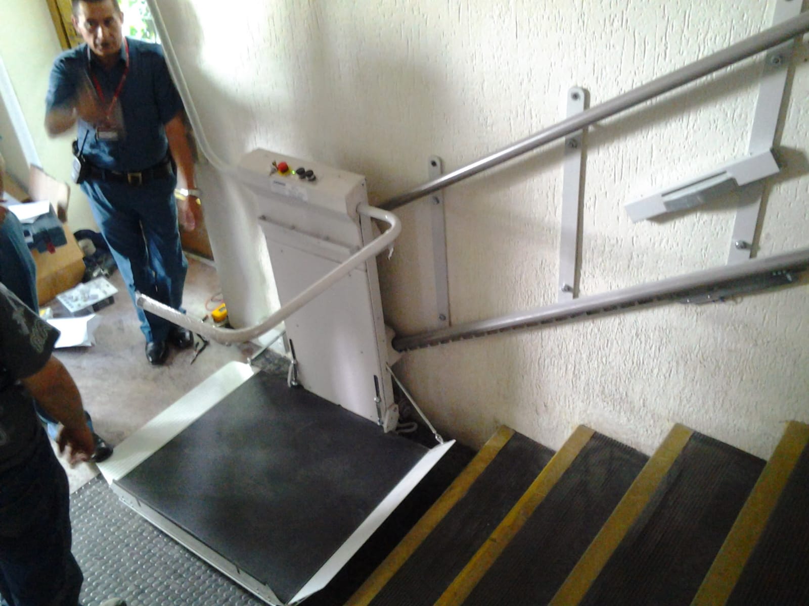 Plataforma elevadora para sillas de ruedas instalada al costado de una escalera, supervisada por técnico de mantenimiento.